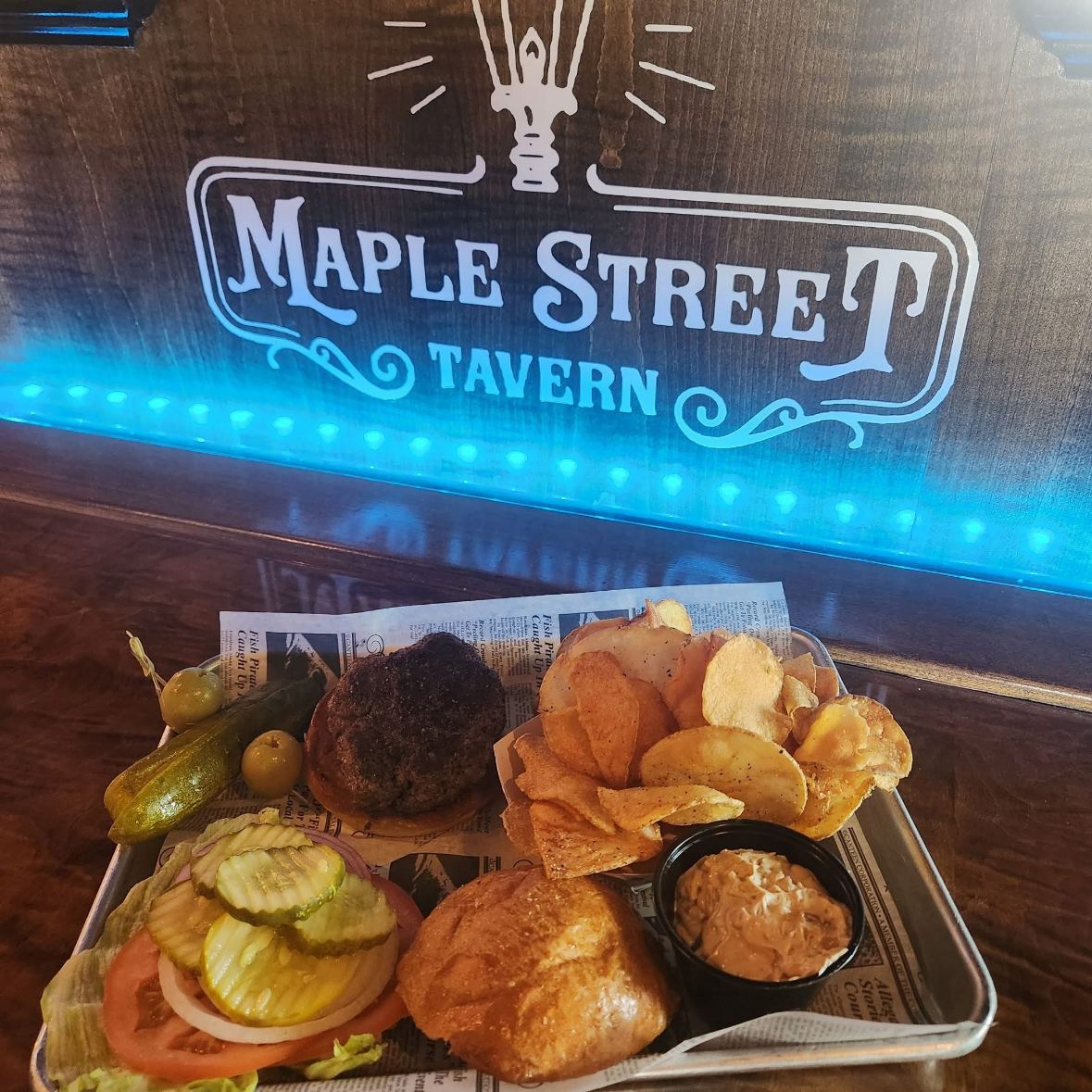 A burger on a tray with homemade chips, pickles and dip under the glowing tavern sign