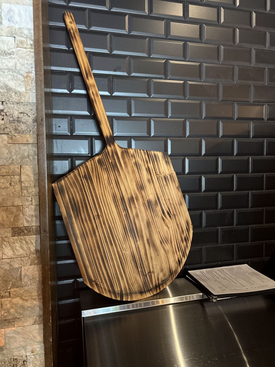 A well-used wooden pizza peel resting against the black brick wall